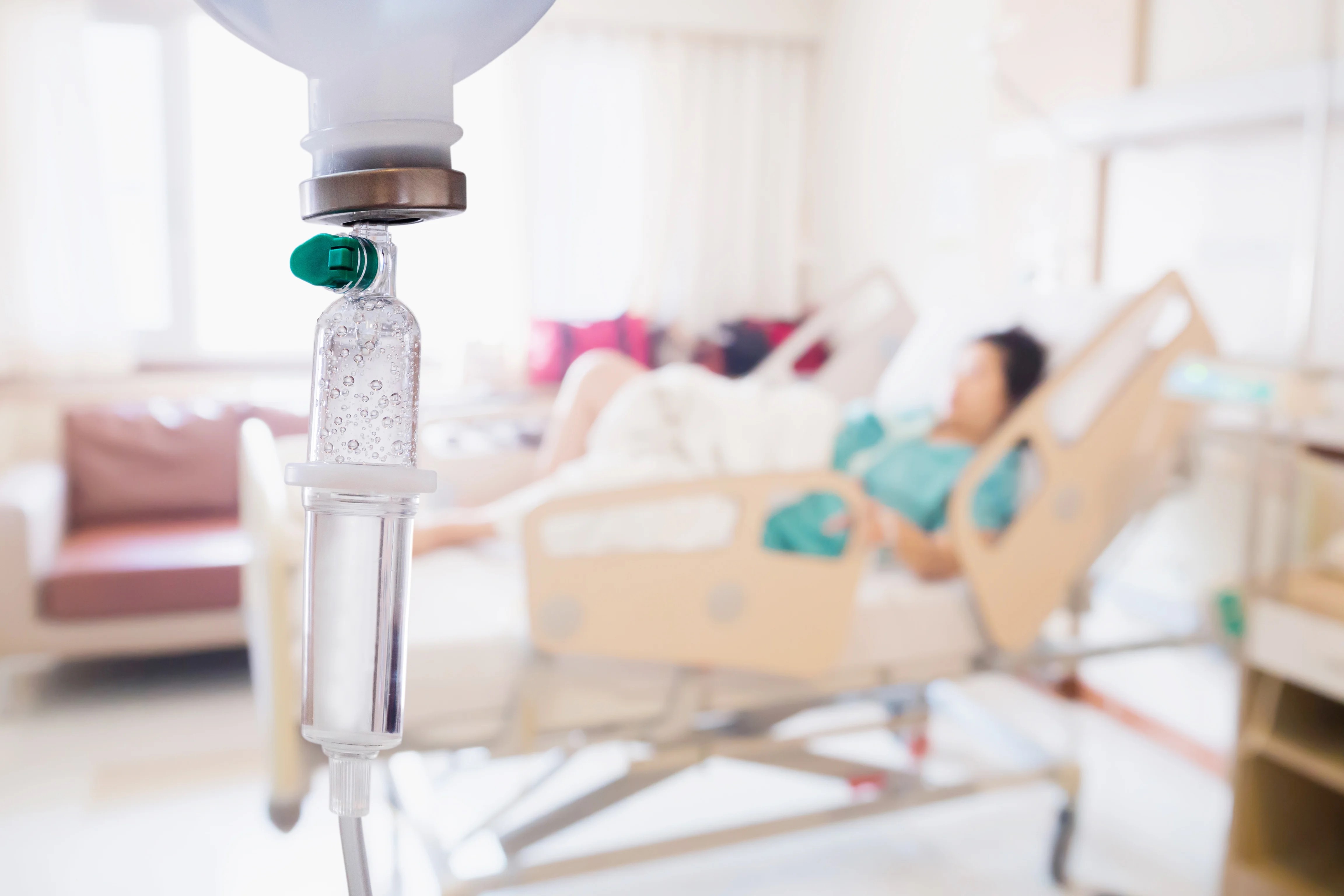 Photo of a woman laying in a hospital bed, with the focus on an IV drip in the foreground