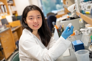 a woman wearing a lab coat in a research lab smiles at the camera as she pipettes liquid from a small tube