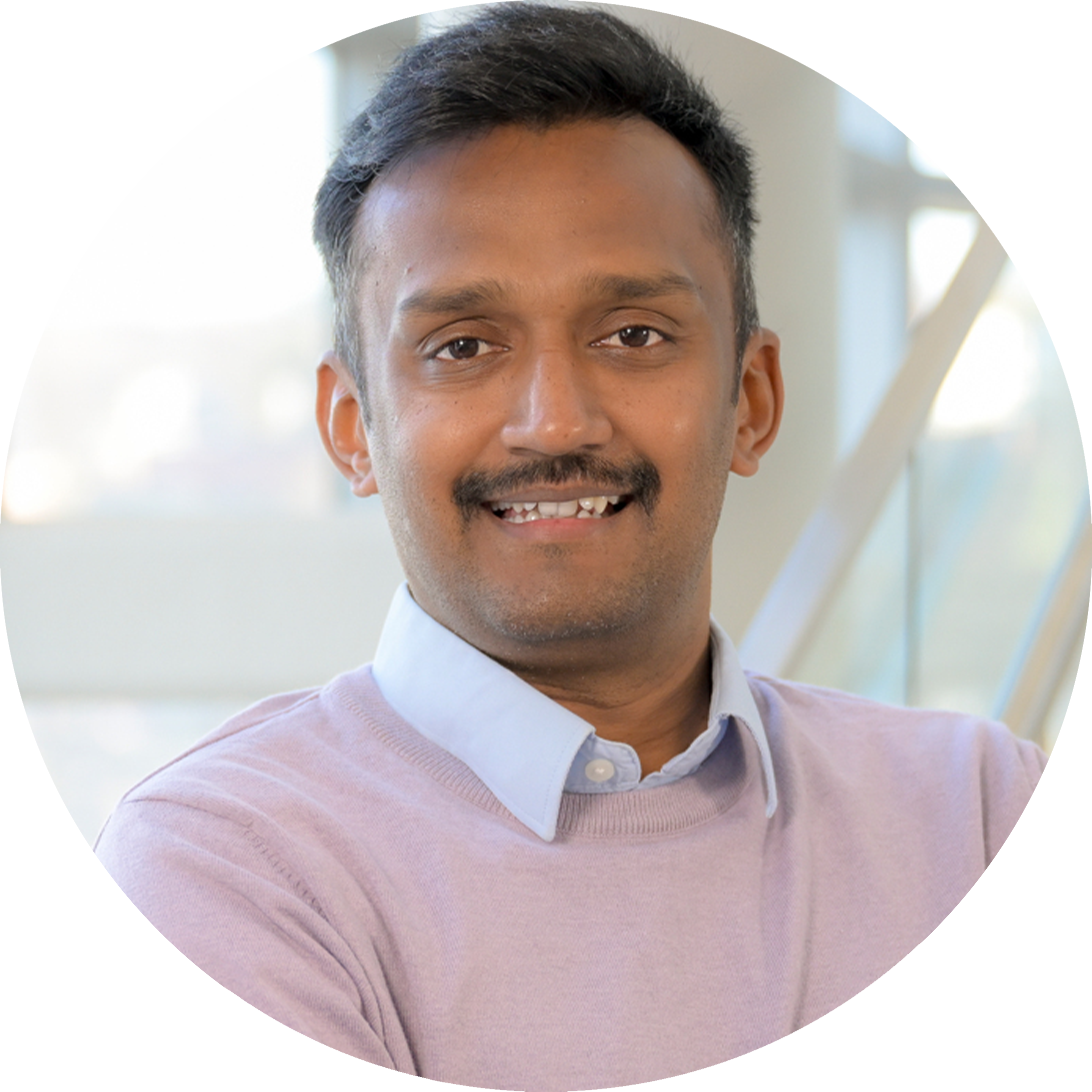 A headshot of Varanasi Siva Karthik, smiling broadly with arms crossed. He is wearing a lavender sweater over a light blue collared shirt, near a staircase with large glass windows in the background.