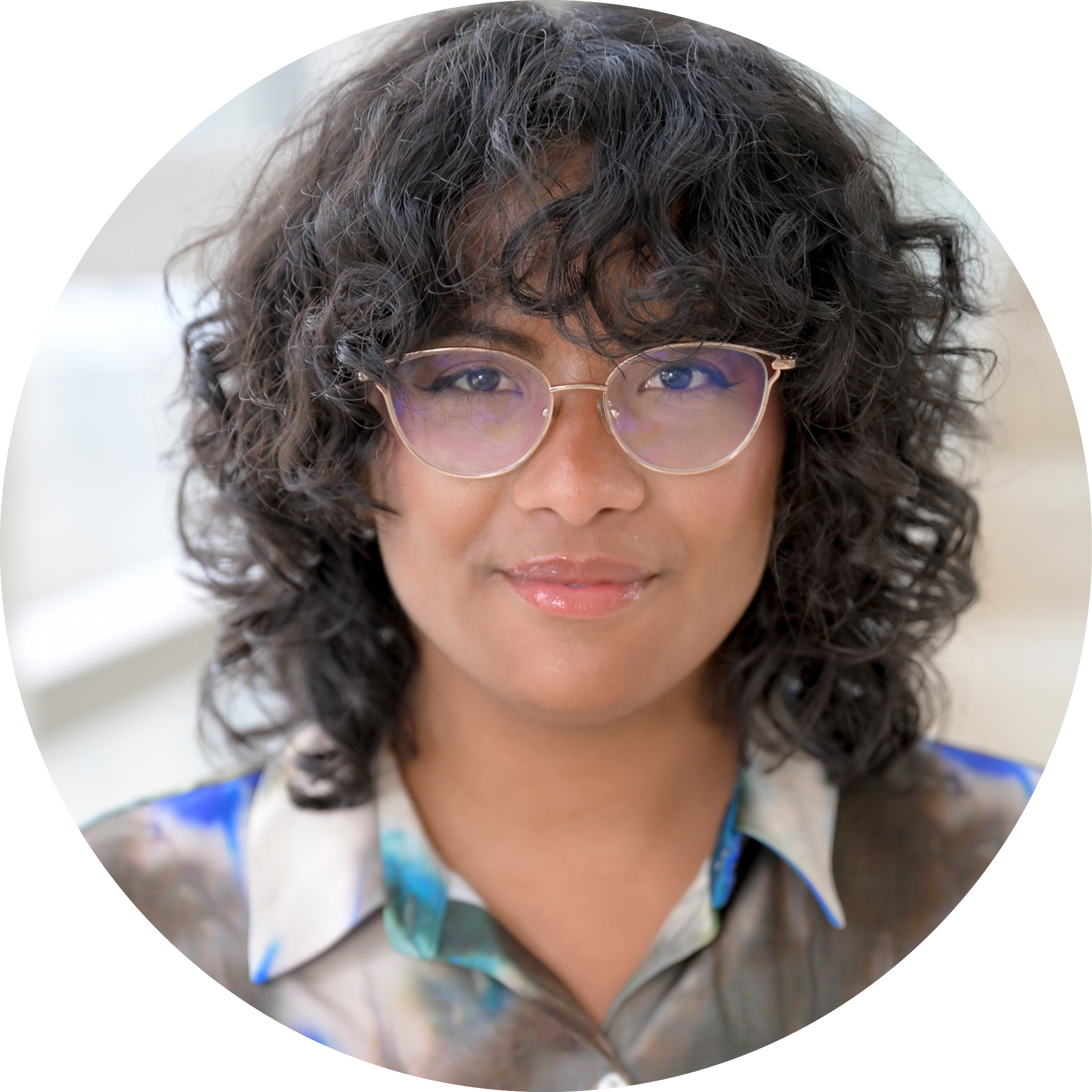 Circular portrait of Natasja Hirabayashi, a mixed-race woman with short-curly brown hair and glasses, wearing a colorful collared shirt