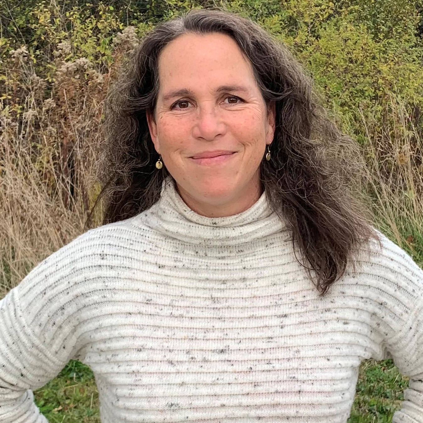 White woman with brown curly hair smiling wearing a white sweater