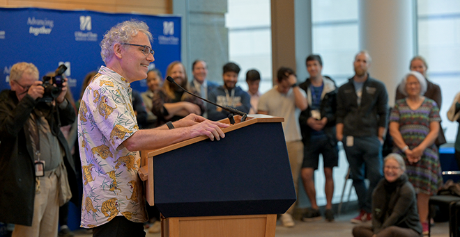 Victor Ambros speaking to reporters at a press conference at UMass Chan Medical School on Oct. 7. In the foreground, Dr. Ambros stands at a podium and is speaking into a microphone. In the background is a sea of people, including reporters who are attending the press conference.