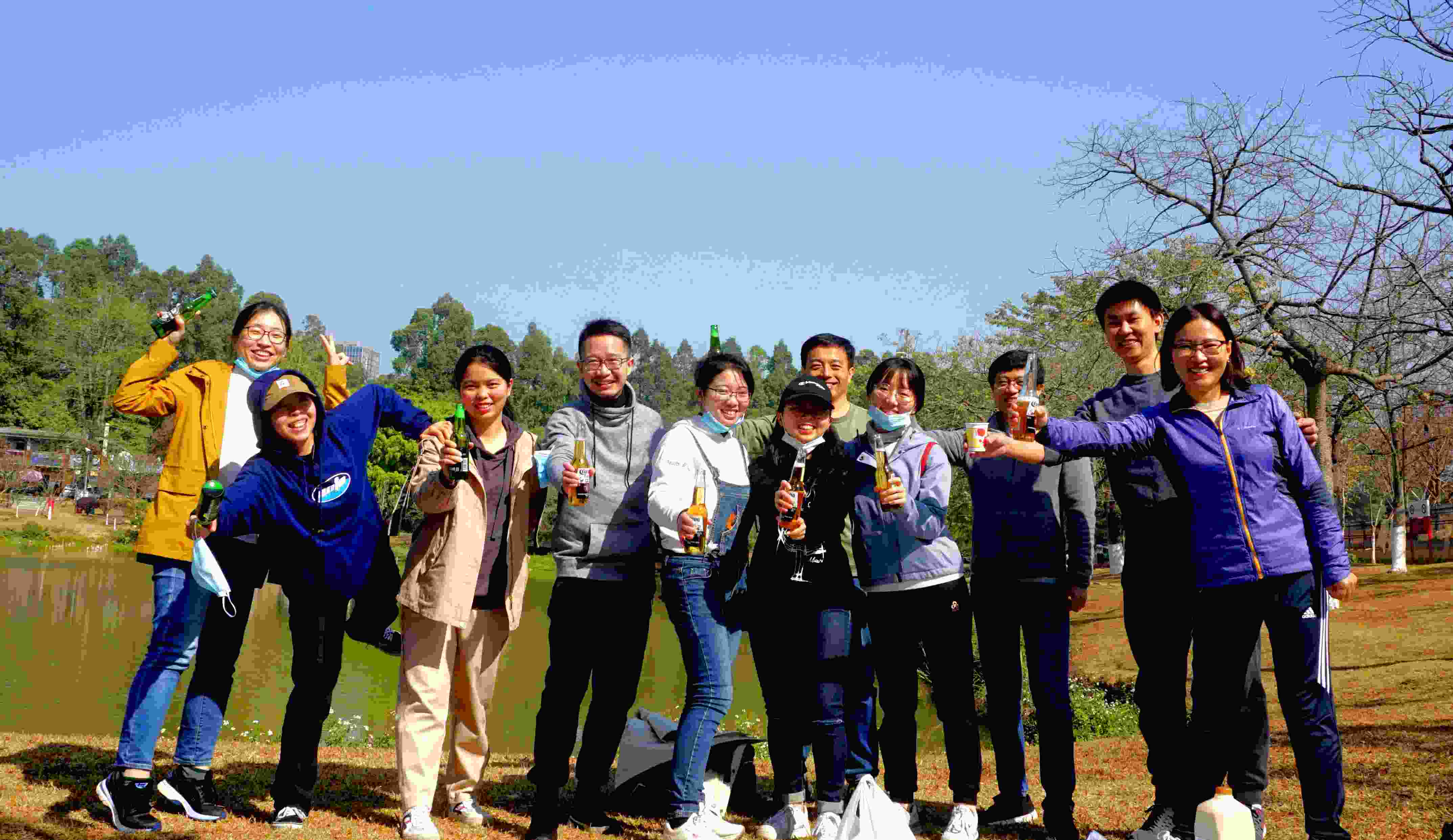 A group of eleven people stand outdoors in front of a lake, smiling and holding up drinks